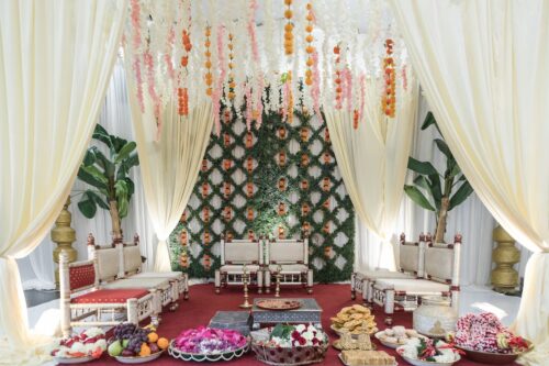 The ceremony space with white curtains, hanging garlands of white, orange, and pink flowers, a latticed background of greenery, low ornate chairs, and platters of fruits, sweets, and flower petals, at a micro-wedding at the Sri Lakshmi Temple in Ashland, MA, photographed by Spagnolo Photography for a micro-wedding coverage package.