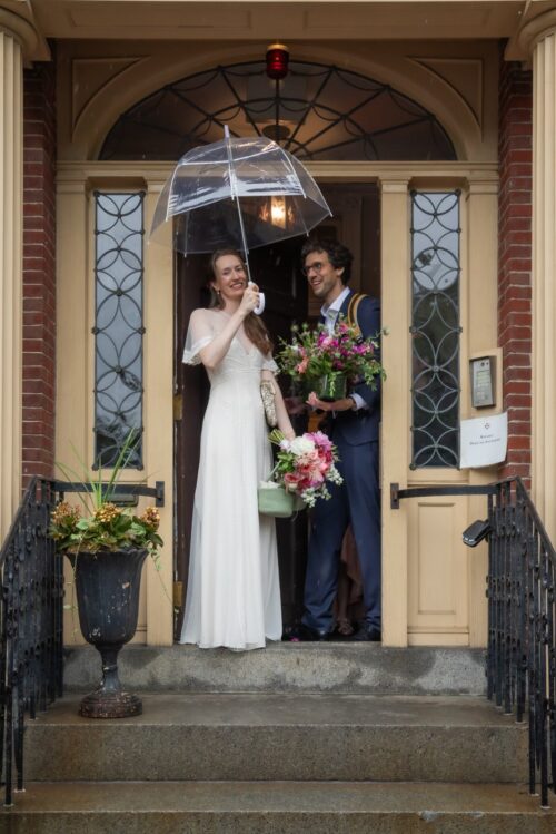 A bride and a groom smile at the camera as they emerge from the yellow entrance of the King's Chapel Parish House in Boston's Beacon Hill where they had their micro wedding