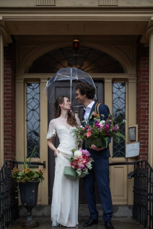 A bride and a groom smile at each other under a clear umbrella in the yellow doorway of the King's Chapel Parish House in Boston's Beacon Hill where they had their micro wedding