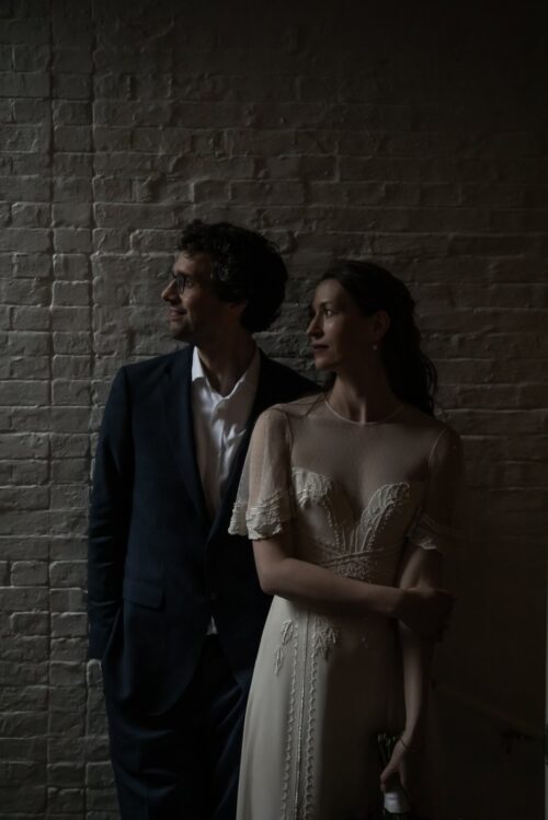 A dramatic and moody Boston wedding portrait of a bride and groom standing in front of a brick wall and looking towards the light, photographed at the historic King's Chapel Parish House during their micro-wedding by Boston wedding photographers Spagnolo Photography.