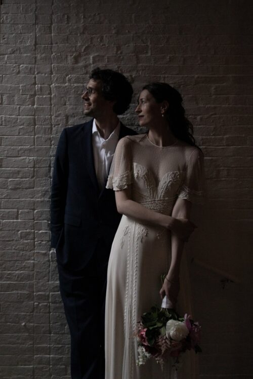 A dramatic and moody Boston wedding portrait of a bride and groom standing in front of a brick wall and looking towards the light, photographed at the historic King's Chapel Parish House during their micro-wedding by Boston wedding photographers Spagnolo Photography.